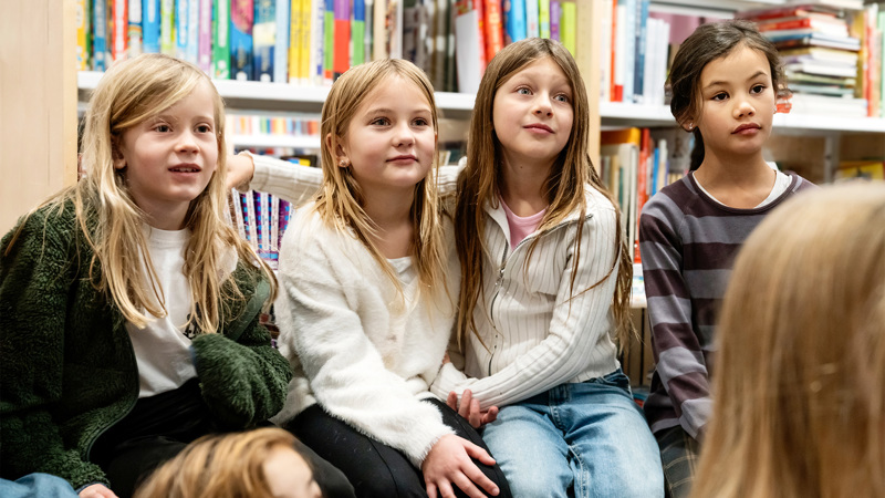 Barn sitter i ett bibliotek och lyssnar på någon som pratar.