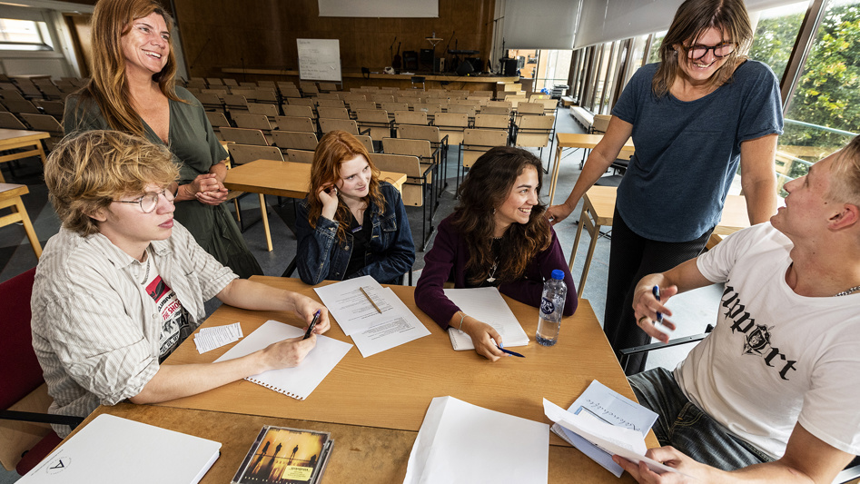 Fyra elever sitter vid ett bord och jobbar med papper och penna. Två lärare står intill.