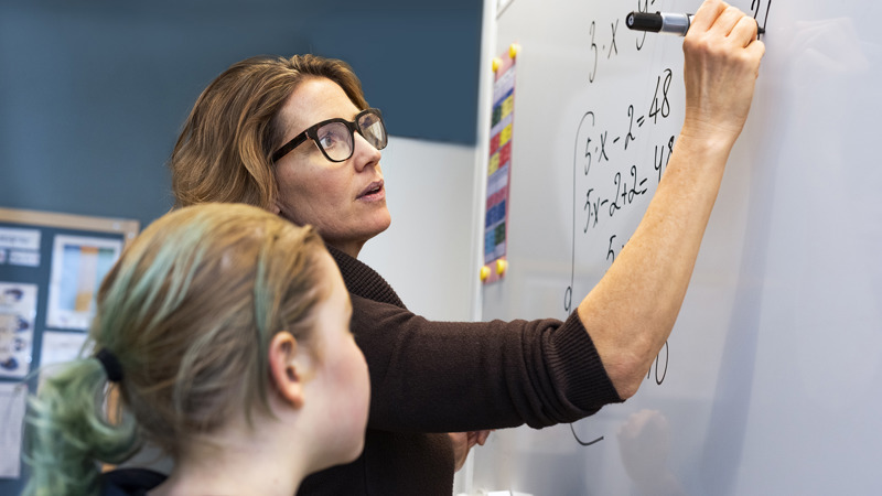En lärare skriver på en whiteboardtavla. En elev tittar på.