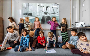 En förskoleklass sitter på golvet i ett klassrum. Bakom dem syns ett stort svartvitt foto.