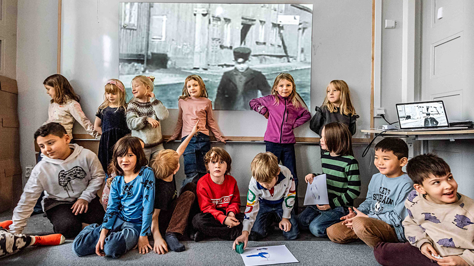 En förskoleklass sitter på golvet i ett klassrum. Bakom dem syns ett stort svartvitt foto.
