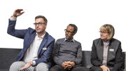 Tre personer sitter på rad i en soffa. Personen till vänster håller ut sina armar och gestikulerar.