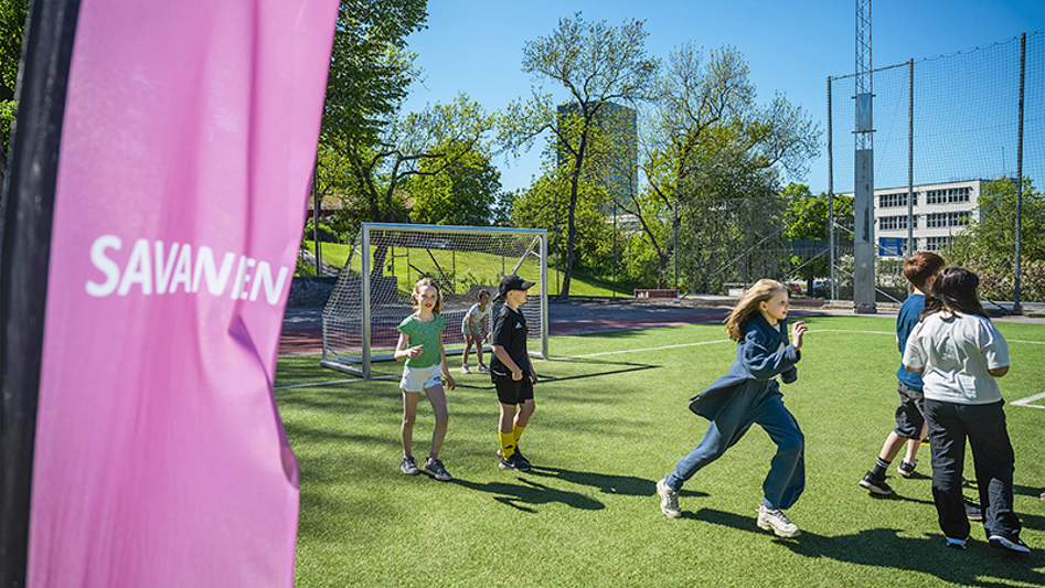 Skolbarn som springer omkring på en gräsbelagd fotbollsplan. Solig och glad känsla.