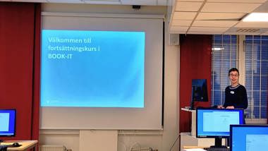 En kvinna står bredvid en skärm längst fram i en datorsal.